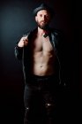 Shirtless cool hipster in hat adjusting leather jacket and looking at camera on black background — Stock Photo