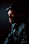 Cool hipster in hat and leather jacket looking away on black background — Stock Photo