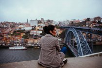 Back view of beautiful female in trendy outfit looking away while sitting near modern bridge in majestic aged city — Stock Photo