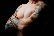 Side view of crop shirtless hipster with crossed hands and tattoos on hands on black background — Stock Photo