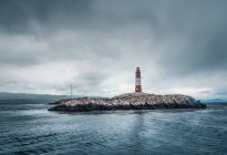 Faro su piccola isola rocciosa nel mare tempestoso il giorno coperto — Foto stock