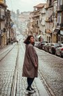 Side view of lovely young lady in stylish coat looking away while standing on paved road on street of ancient city — Stock Photo