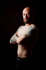 Vista laterale di hipster serio calvo senza maglietta con tatuaggi sulle mani che guardano la fotocamera su sfondo nero — Foto stock