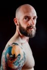 Side view of shirtless bald serious hipster with tattoos on hands looking at camera on black background — Stock Photo