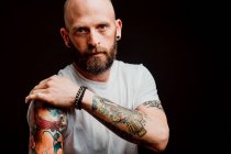 Hipster sin pelo barbudo en camiseta que muestra tatuajes en las manos sobre fondo negro - foto de stock
