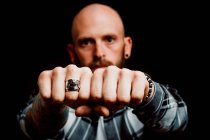 Bearded serious hipster in shirt with tattoos on hand showing fists with ring on black background — Stock Photo
