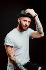 Joyeux hipster barbu en chapeau et t-shirt avec des tatouages sur les bras sur fond noir — Photo de stock