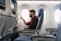 Vista lateral do cara barbudo que navega o gadget da aeronave ao sentar-se no assento confortável dentro do avião moderno — Fotografia de Stock