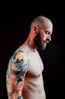 Vue latérale de hipster sérieux chauve torse nu avec des tatouages sur les bras regardant loin sur fond noir — Photo de stock