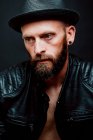 Cool hipster in hat and leather jacket looking away on black background — Stock Photo
