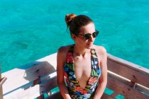 Dama en traje de baño y gafas de sol sentada en el asiento cerca del mar azul en Jamaica - foto de stock