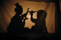 Silhouette nera di bambino e donna che giocano con dinosauri giocattolo seduti alla luce della lampada dietro le lenzuola a casa — Foto stock