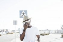Homme noir branché dans le chapeau et les lunettes de soleil debout dans la lumière vive sur la rue en regardant la caméra — Photo de stock