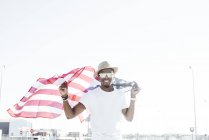 Content stylish black man in sunglasses and hat holding American flag smiling at camera in sunlight — Stock Photo