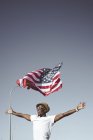 Excited black man in sunglasses and hat holding hands apart with American flag flying above head on blue sky — Stock Photo