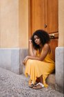 Gorgeous black woman in dress sitting on porch on street and looking at camera — Stock Photo
