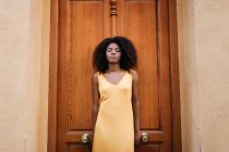 Мечтательная чёрная женщина в жёлтом платье прислонилась к двери на улице — стоковое фото