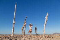 Jovem mulher atraente em pé na postura asana entre troncos secos na praia em dia ensolarado — Fotografia de Stock