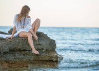 Sogno ad occhi aperti giovane donna in beachwear seduta sulla pietra al mare e guardando la fotocamera — Foto stock