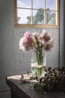 Деревянный стол с букетом розовых хризантем в вазе между упавшими лепестками и белой стеной с окном — стоковое фото