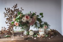 Dish with tasty cake decorated bloom bud on wooden table with bunch of chrysanthemums, roses and plant twigs in vase between dry leaves on grey background — Stock Photo