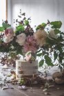 Gericht mit leckerem Kuchen dekoriert Blütenknospe auf Holztisch mit Bund Chrysanthemen, Rosen und Pflanzenzweigen in Vase zwischen trockenen Blättern auf grauem Hintergrund — Stockfoto