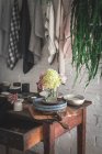 Деревянный стол со свежими розовыми хризантемами и белыми гортензиями в вазе между сковородкой и кухонной утварью рядом с посудой, висящей на твисте с булавками — стоковое фото