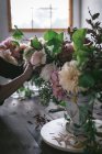 Женщина делает букет из сухих и свежих роз, хризантем и веток растений в ретро-вазе на деревянной доске на сером фоне — стоковое фото
