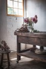 Tavolo in legno con stoviglie e mazzi di fiori freschi in vasi con acqua vicino alla parete bianca — Foto stock