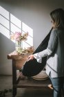 Vista lateral da senhora segurando caso perto de mesa de madeira com buquê de crisântemos rosa em vaso com água entre pétalas caídas perto da parede branca com sol — Fotografia de Stock