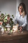 Side view of lady placing dish with tasty cake decorated bloom bud on wooden table with bunch of chrysanthemums, roses and plant twigs in vase between dry leaves on grey background — Stock Photo