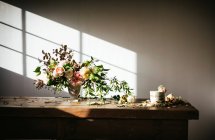 Plato con sabroso pastel decorado brote de flor en la mesa de madera con racimo de crisantemos, rosas y ramitas de plantas en jarrón entre hojas secas sobre fondo gris - foto de stock