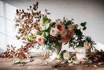 Plato con sabroso pastel decorado brote de flor en la mesa de madera con racimo de crisantemos, rosas y ramitas de plantas en jarrón entre hojas secas sobre fondo gris - foto de stock