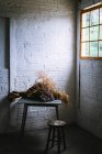 Concept of bouquet of dry coniferous twigs in craft paper on table near stool in grey murk room with brick walls — Stock Photo