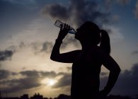 Silhouette of lady in sportswear drinking water from bottle on wonderful heaven background with clouds in evening — Stock Photo