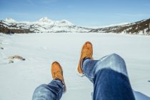 Cultivo piernas masculinas en jeans y botas de invierno sentado en la nieve en el día soleado cerca de las colinas en Cerdanya, Francia - foto de stock