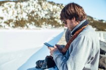 Vista lateral del joven fotógrafo con cámara profesional y en el teléfono móvil entre montañas en la nieve en Cerdanya, Francia - foto de stock