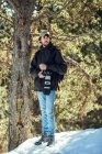 Jeune homme en lunettes de soleil et casquette avec sac à dos regardant loin tout en tenant une caméra professionnelle entre forêt d'hiver à Cerdanya, France — Photo de stock