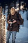 Junger Mann mit Sonnenbrille und Schirmmütze mit Rucksack blickt auf professionellen Kamerabildschirm zwischen Winterwald in Cerdanya, Frankreich — Stockfoto