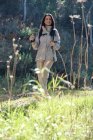 Весела леді в пальто, що йде з пішохідними палицями на пішохідній доріжці в лісі в сонячний день — стокове фото