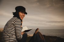 Vue arrière de la femme élégante dans le volume de lecture de chapeau et assis sur la prairie près de la balise sur la rive et le ciel merveilleux — Photo de stock