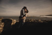 Vue latérale d'élégant couple élégant embrasser près des rochers sur la côte près de la mer et le ciel merveilleux — Photo de stock