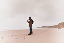 Vista lateral del tipo en ropa de abrigo con la mochila fumar pipa en la costa de arena cerca del mar y el cielo en las nubes - foto de stock