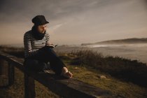 Elegante mujer en gorra tomando notas en bloc de notas y sentada en el asiento en la costa cerca del mar ondulante - foto de stock