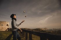 Элегантная женщина в шапке жонглирует мячами на траве у берегов моря и неба с солнцем — стоковое фото