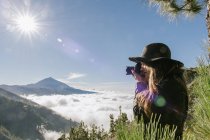 Vista lateral do fotógrafo feminino em pé no topo da colina tirando fotos na paisagem nublada no dia ensolarado — Fotografia de Stock
