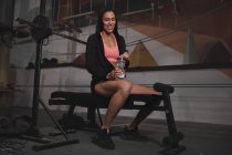Юная стройная веселая женщина в спортивной одежде с бутылкой воды, сидящей на скамейке в спортзале — стоковое фото