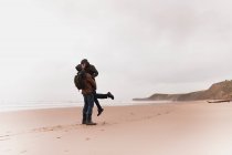 Side view of young man with backpack in warm wear holding on hands and kissing woman on sand beach near sea and hills — Stock Photo