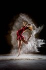 Вид збоку молодої тонкої балерини в червоній сукні збільшуючи ногу і руки між туманом в темній кімнаті — стокове фото