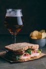 Delicious sandwich with ham, cheese and greens with glass of beer and chips on dark background — Stock Photo